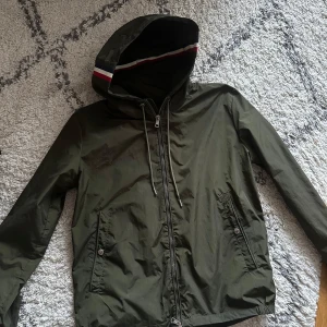 Olivgrön vindjacka från Moncler - Snygg olivgrön vindjacka från Moncler med huva och dragkedja framtill. Jackan har två fickor med knappar och Moncler-logga på ärmen. Perfekt för dig som vill ha en stilren och sportig look. Priset är 1799kr eller högsta bud. Först till kvarn som gäller. Pm för mer info. 
