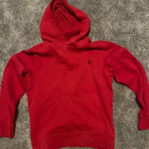 Röd hoodie från Polo Ralph Lauren - Röd hoodie från Polo Ralph Lauren med klassisk broderad logga på bröstet. Tröjan har huva, magficka och ribbade muddar vid ärmslut och nederkant. Perfekt för en avslappnad och sportig stil.