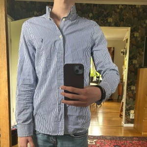 Blåvit randig skjorta från Ralph Lauren - Klassisk blåvit randig skjorta från Ralph Lauren med knappar framtill och liten broderad logga på bröstet. Skjortan har lång ärm och en smal passform som ger en stilren look. Perfekt till jeans eller chinos. Storlek 18 motsvarar storlek S. nypris runt 1500