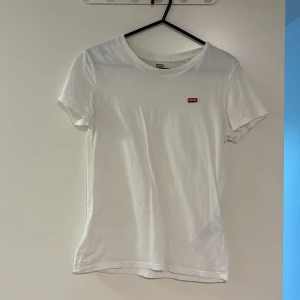 Vit t-shirt från Levi's - Klassisk vit t-shirt från Levi's med liten röd logotyp på bröstet. Modellen är enkel med rund halsringning och korta ärmar. Perfekt basplagg i mjuk bomull som passar till allt.
