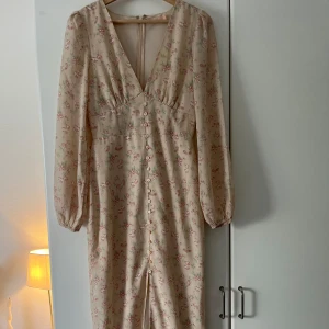 Blommig långklänning med v-ringning - Romantisk långklänning i ljusbeige med blommigt mönster i rosa och grönt. Djup v-ringning, långa ärmar och knappar framtill. Klänningen har en feminin siluett och lätt puffade axlar.🩷