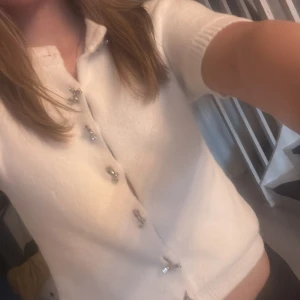 Vit croppad kofta med dekorativa knappar - Säljer en söt vit croppad kofta med korta ärmar och glittriga dekorativa knappar framtill. Perfekt för dig som gillar en stilren men ändå unik look. Koftan har en mjuk känsla och passar till många outfits.