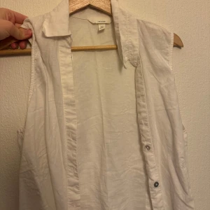 Vit ärmlös skjorta  - Säljer en stilren vit ärmlös skjorta från H&M. Skjortan har klassisk krage och knappar framtill. Perfekt att styla lager på lager eller bära som den är under varma dagar. I storlek S men passar även M beroende på hur man vill att den ska sitta