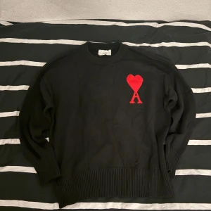 Svart stickad tröja från Ami Paris - Svart stickad tröja från Ami Paris med rund halsringning och ett stort rött hjärta med bokstaven A broderat på bröstet. Tröjan har långa ärmar och bred ribbstickning vid ärmslut och nederkant. Perfekt för dig som gillar stilrena plagg med en twist. Kontakta för mer information.