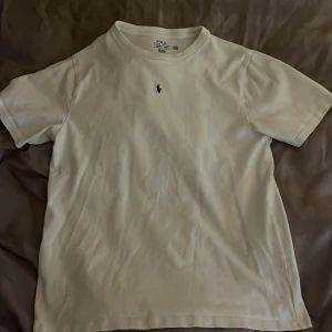 Vit t-shirt från Polo Ralph Lauren - Klassisk vit t-shirt från Polo Ralph Lauren med mörkblå broderad logga på bröstet. T-shirten har rund hals och korta ärmar, perfekt för en clean och stilren look i sommar