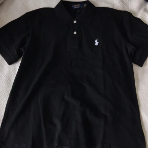 Svart pikétröja från Polo Ralph Lauren - Klassisk svart pikétröja från Polo Ralph Lauren med vit broderad logga på bröstet. Tröjan har korta ärmar, krage och knäppning med två knappar. Modellen är custom slim fit och tillverkad i bomull.