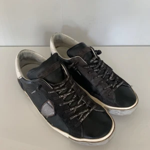 Svarta sneakers från Philipp Plain, 42 - Säljer ett par svarta Philippe Model sneakers med vita detaljer och logga på sidan. Skorna har snörning med text på skosnörena och en platt sula. Perfekta för dig som gillar stilrena och sportiga skor. Sparsamt använda.