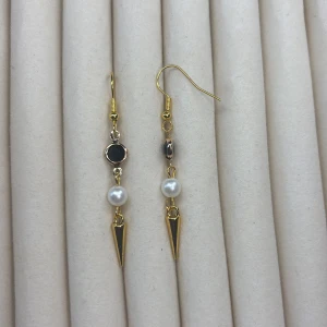Guldiga örhängen med pärlor och svarta detaljer - Säljer ett par unika guldiga örhängen med svart sten, vit pärla och spetsig hängande detalj. Perfekta för dig som vill sticka ut med en cool accessoar. Säljs som par.