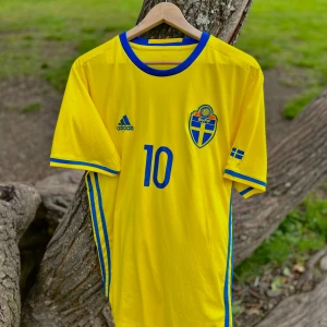 Sverige Hemmatröja 2016 - Gul fotbollströja från Adidas med blå detaljer, svenska landslagets emblem och nummer 10 på bröstet. Med namnet 'Ibrahimović' på ryggen. Klassisk design med blå ränder på sidorna och korta ärmar.