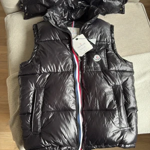 Moncler väst - Snygg svart dunväst från Moncler med glansig finish och klassisk logotyp på bröstet. Luva som går att ta av och sätta på, pris kan sänkas vid snabb affär. Funkar till kyliga dagar och varmare med bara t shirt under