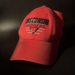 Röd keps Wisconsin Badgers - Röd keps med broderad text och logga för Wisconsin Badgers framtill. Justerbar baktill för bra passform. Perfekt för dig som vill visa ditt stöd för laget.