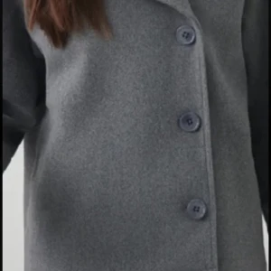 Grå kappa med knappar - Stilren grå kappa med stora knappar framtill. Klassisk rak modell som passar perfekt till kyligare dagar. Enkel design utan synliga detaljer förutom knapparna. Perfekt för dig som gillar en tidlös look.