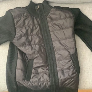 Svart quiltad jacka med stickade ärmar - Säljer en svart jacka med quiltad framsida och stickade ärmar samt rygg. Jackan har dragkedja framtill och två fickor. Perfekt för kyliga dagar när du vill ha både stil och komfort. Har använda typ 2 gånger först man till kvarn 