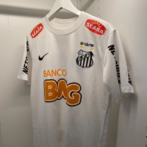 Vit Santos fotbollströja Neymar Jr - Säljer en vit Santos fotbollströja med Neymar Jr och nummer 11 på ryggen. Tröjan har svarta tryck, sponsorloggor och klubbmärke på bröstet. Kortärmad modell i lätt material, perfekt för match eller träning. Den är i riktigt bra skick och 100% äkta. 