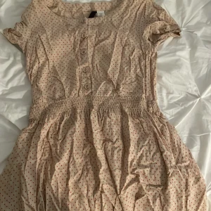 Beige kortklänning med röda prickar och knappar - Söt beige kortklänning med röda prickar och knappar framtill. Klänningen har korta ärmar, markerad midja med resår och ett lätt, luftigt tyg. Perfekt för varma dagar och enkel att styla med sneakers eller sandaler.