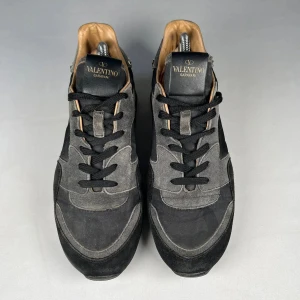 Valentino Rockrunner sneakers i svart och grå mocka - Säljer ett par Valentino Rockrunner sneakers i svart och grå mocka med svarta snören och ikoniska nitar runt sulan. Skorna har en sportig design med rund tå och platt sula. Perfekta för dig som vill ha en lyxig touch på vardagslooken.