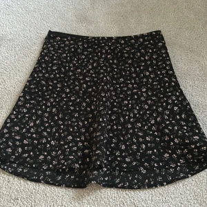 Svart blommig kjol från H&M Divided - Svart kjol från H&M Divided med små blommiga mönster i vitt och rosa. Kjolen har en lätt utsvängd form och är kort i modellen. Perfekt för dig som gillar en söt och enkel stil.