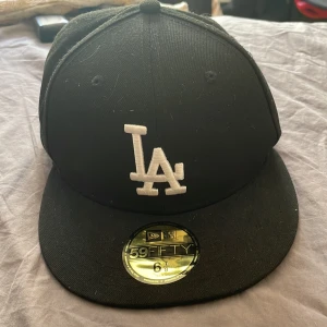 Svart LA-keps från New Era 59FIFTY - Svart keps från New Era 59FIFTY med LA-logga broderad framtill i vitt. Klassisk flat peak och äkta MLB-merchandise. Tillverkad i polyester och passar huvudomkrets 54,9 cm.