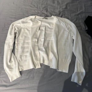 Vit kofta i bomull - Säljer en klassisk vit kofta med långa ärmar och knäppning framtill. Koftan är tunn och har ribbade muddar vid ärmslut och nederkant. Perfekt att slänga över en topp för en enkel och stilren look.