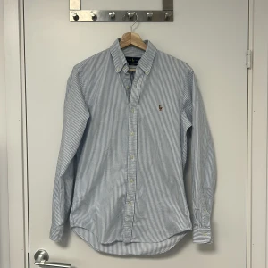 Ralph Lauren Oxford Skjorta - Klassiska Oxford skjortan från Ralph Lauren i den ikoniska blå-randiga färgen. Skjortan har den ikoniska multi-färgade broderade loggan på bröstet. Hör av er vid funderingar!