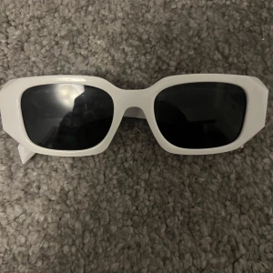 Vita solglasögon från Prada - Snygga vita solglasögon från Prada med rektangulära bågar och svarta glas. Klassisk design med guldiga Prada-loggor på sidorna. Perfekt accessoar för att lyfta din stil.