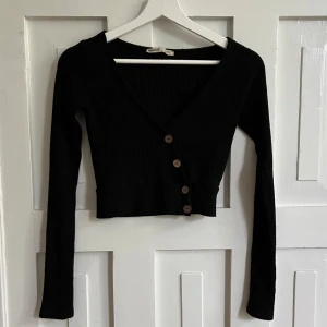 Svart ribbad kofta från Pull&Bear - Säljer en svart croppad kofta från Pull&Bear. Tröjan är ribbad, har v-ringning och har fyra knappar framtill som är fejk knappar så dom går inte att knäppa upp.