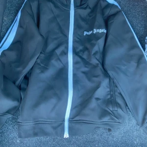 Svart kofta från Palm Angels - Palm Angels svart kofta med vita ränder längs ärmarna och vit dragkedja framtill. Jackan har ståkrage och logga tryckt på bröstet. Perfekt för en sportig och trendig look.