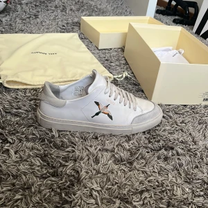 Vita sneakers med broderad fågel från Axel Arigato - Säljer ett par vita sneakers från Axel Arigato med en snygg broderad fågel på sidan. Skorna har snörning och rund tå, och är tillverkade i skinn. Perfekta för dig som gillar stilrena och trendiga sneakers.