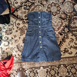 Jeansklänning med knappar - Säljer en snygg axelbandslös jeansklänning med knappar framtill och fickor. Klänningen har en markerad midja och är i klassisk blå denim. Perfekt för dig som gillar en cool och avslappnad stil.