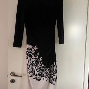 Svart och vit blommig långklänning från Lauren Ralph Lauren - Elegant vintage midiklänning från Lauren Ralph Lauren i svart och vitt med blommigt mönster nertill. Klänningen har lång ärm och är tillverkad i stretchig polyesterblandning. Perfekt för dig som vill ha en stilren och klassisk look. 