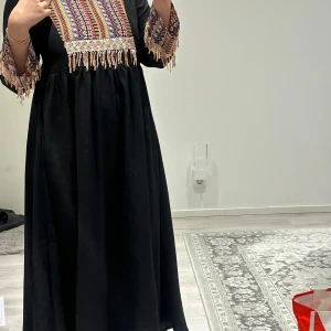 Svart långklänning med broderad överdel och fransar - Svart långklänning med färgglad broderad överdel och ärmslut i rött, blått, gult och vitt. Klänningen har dekorativa fransar och paljettdetaljer vid bröstet och ärmarna. 