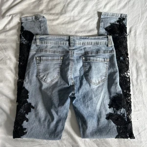 Ljusblå jeans med svarta spetsdetaljer - Unika ljusblå jeans med svarta spetsapplikationer längs båda benen från Realty. Klassisk femficksmodell med normal passform och knappgylf. Byxorna är i strl 38