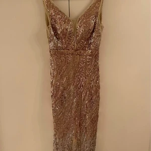 Glittrig champagnefärgad långklänning med v-ringning - Säljer en elegant långklänning i champagnefärg med glittriga paljetter och djup v-ringning. Klänningen har smal passform och är ärmlös, perfekt för fest eller bal. Materialet är syntet och paljetter för extra glamour.