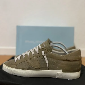 Sneakers från Philippe Model - Snygga beige gröna sneakers från Philippe Model med vita snören och vit platt sula. Skorna har en rund tå och är tillverkade i skinn med diskreta sömdetaljer på sidan. Perfekta för en stilren och avslappnad look. Skorna är sparsamt använda och i storlek 44. Bra skick! Nypris runt 3000 kr.