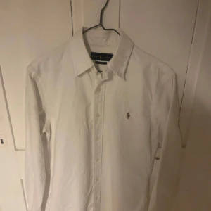 Vit skjorta från Ralph Lauren - Klassisk vit skjorta från Ralph Lauren i slim fit-modell. Skjortan har långa ärmar, knappar framtill och den ikoniska broderade loggan på bröstet. Perfekt för en stilren och tidlös look.