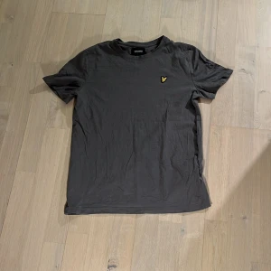 Mörkgrå t-shirt från Lyle & Scott - Säljer en stilren mörkgrå t-shirt från Lyle & Scott med klassisk rund hals och gul broderad logga på bröstet. Perfekt basplagg med normal passform och korta ärmar. Skön och mjuk bomullskvalitet.