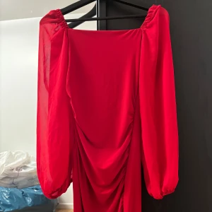 Röd långklänning från Keep Look - Säljer en röd långklänning från New Look med puffiga långärmar och draperad front. Klänningen har en fyrkantig ringning och är tillverkad i ett lätt, syntetiskt material. Perfekt för dig som vill sticka ut med en färgstark look!