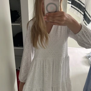 Vit broderad klänning med v-ringning - Superfin vit klänning med v-ringning och långa ärmar. Klänningen har broderade detaljer och volanger i kjolen, vilket ger en romantisk känsla. Perfekt för sommarens alla tillfällen!