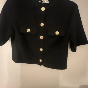 Svart cardigan jacka med guldknappar - Stilren svart cardigan jacka med kort ärm och dekorativa guldknappar framtill. Två fickor med lock och knappdetalj på bröstet. Perfekt för en trendig och elegant look.