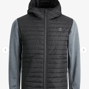 Svart hybridjacka med huva från Jack and Jones - Snygg svart hybridjacka från Jack and Jones  med quiltad framsida och huva. Ärmarna är grå och i mjukare material, medan kroppen är vadderad och har dragkedja hela vägen. Perfekt för lager-på-lager och stilren look.
