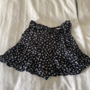 Svart blommig kortkjol från Zara - Supersöt svart kortkjol från Zara med vitt blommönster och volangdetaljer. Kjolen har omlottmodell med knyt i midjan och är perfekt för dig som gillar en feminin och trendig stil.