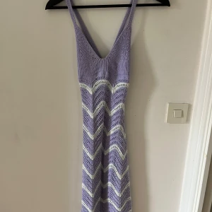 Ljuslila strand klänning med mönster - Superfin långklänning i ljuslila stickat material med vitt vågigt mönster. Djup v-ringning och smala axelband, perfekt för varma dagar. Klänningen har en figurnära passform och är både mjuk och bekväm.