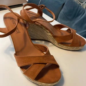 Bruna kilklackade sandaler från Esmara - Snygga sandaler med kilklack i flätad jute och bruna remmar i mockaimitation. Skorna har öppen tå och justerbart spänne runt vristen. Perfekta för varma dagar och ger en somrig känsla till outfiten.
