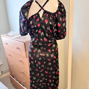 Svart blommig klänning med slits - Säljer en svart klänning med färgglatt blommönster i rött, grönt och gult. Klänningen har puffiga kortärmar och en söt knytning vid halsen. Den har en slits som ger en extra touch. Perfekt för en somrig look!