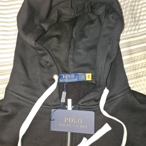 Svart hoodie från Polo Ralph Lauren - Säljer en svart hoodie från Polo Ralph Lauren med dragkedja och vit snörning i huvan. Klassisk röd logga broderad på bröstet och två fickor framtill. Perfekt för en stilren och avslappnad look.