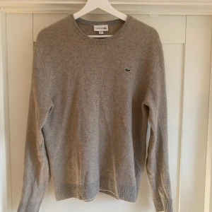 Grå stickad tröja från Lacoste - Säljer en klassisk grå stickad tröja från Lacoste med rund halsringning och den ikoniska gröna krokodilloggan på bröstet. Tröjan är tillverkad i en mjuk blandning av ull, viskos, polyamid, silke och kashmir. Perfekt för lager-på-lager under kyliga dagar.