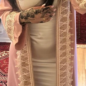 Beige klänning med rosa spetsdetaljer - Elegant beige klänning med vackra rosa spetsdetaljer längs kanterna. Klänningen har en figurnära passform och långa ärmar. Perfekt för speciella tillfällen.