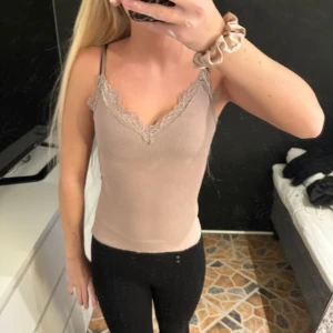 Beige ribbad linnetopp med spetsdetaljer - Säljer en stilren beige ribbad linnetopp med vacker spetskant vid den v-ringade halsen. Toppen har smala axelband och en tight passform, perfekt för en chic och bekväm look.