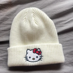 Vit ribbad mössa med Hello Kitty - Söt vit ribbad mössa med en broderad Hello Kitty på framsidan. Perfekt för att hålla sig varm och stilren under kyliga dagar. Den har en uppvikt kant och en ikonisk röd rosett på Hello Kittys huvud.