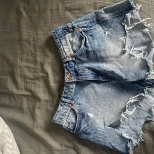 Blå jeansshorts med fransar - Säljer ett par coola blå jeansshorts med fransiga kanter och slitna detaljer. Shortsen har en klassisk femficksdesign och knappgylf. Perfekta för en avslappnad sommarlook!